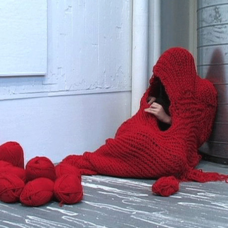 Color photograph of a person crouched on ground almost completely enclosed in a bright red crocheted cocoon. You can only see the hand of the person continuing to work on closing up the opening. There are balls of red yarn in a pile to the left on the grey and white rustic floor